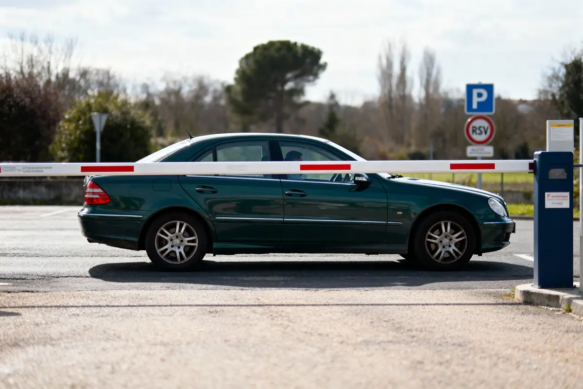 Voiture verte bloquée par une barrière de parking, signalisation en arrière-plan, ciel partiellement nuageux.