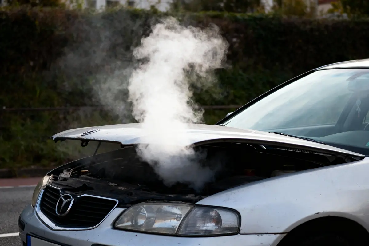 Voiture blanche avec capot ouvert, fumée épaisse s'échappant du moteur, en panne en bord de route.