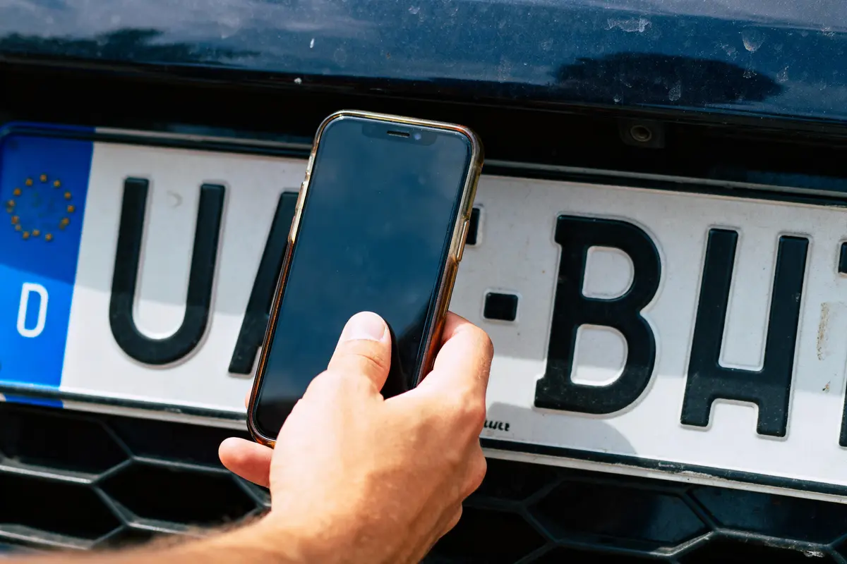 Main tenant un smartphone devant une plaque d'immatriculation européenne d'une voiture.