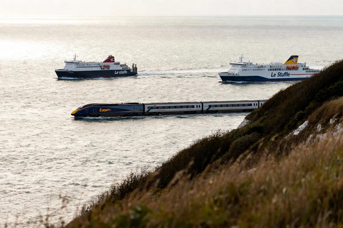 Deux ferries et un train circulant sur l'eau près d'une côte herbeuse, horizon marin en arrière-plan.