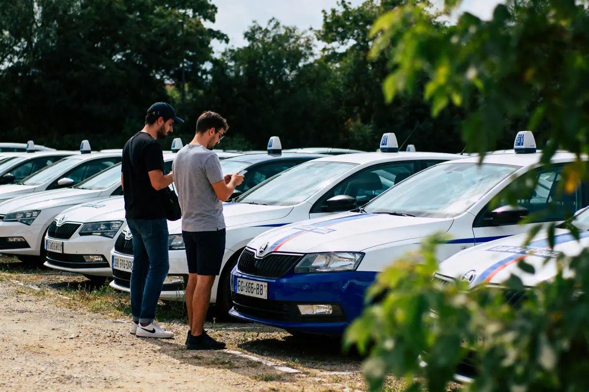Deux hommes debout près de voitures d'auto-école alignées, lisant un document, arbres en arrière-plan.