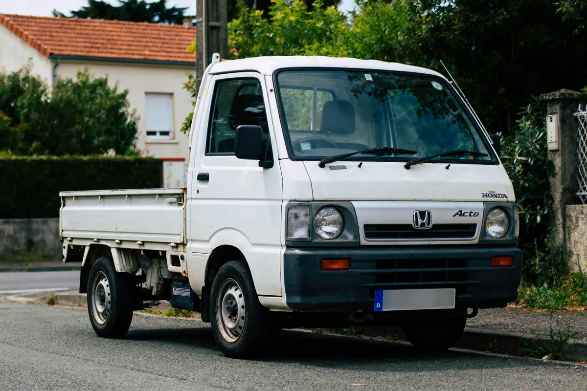 Camionnette blanche Honda Acty stationnée dans une rue résidentielle, devant un bâtiment avec des arbres.
