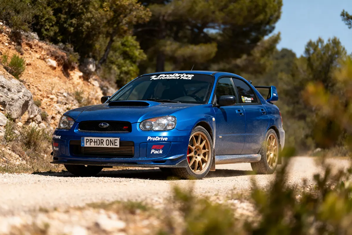 Voiture de rallye bleue Subaru sur chemin de terre, arbres en arrière-plan, plaque d’immatriculation personnalisée.