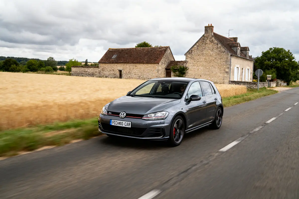 Voiture Volkswagen grise roulant sur route de campagne avec maisons et champs de blé.