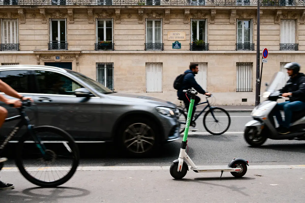 Trottinette, voitures, vélos et scooter en mouvement devant un immeuble parisien. Scène urbaine animée.