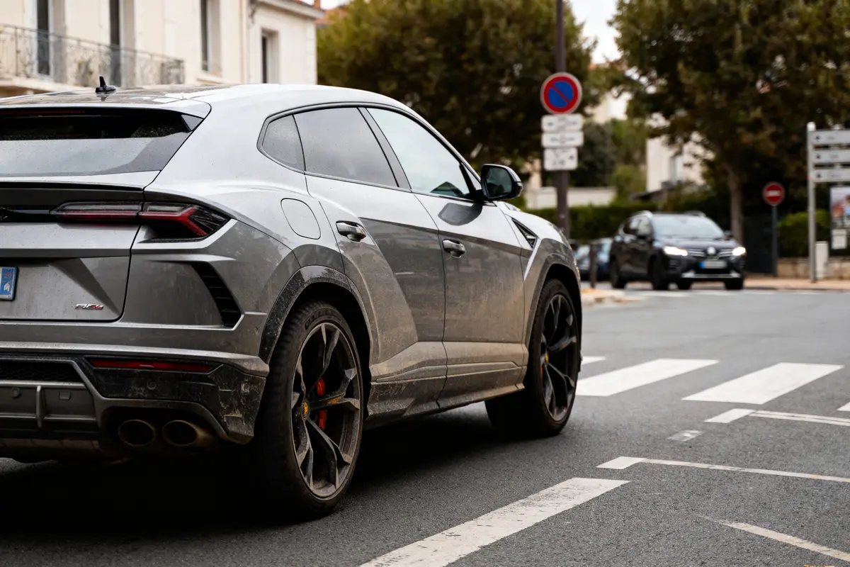 SUV gris sale roulant en ville, passages piétons, arbres, bâtiments en arrière-plan, voitures en circulation.