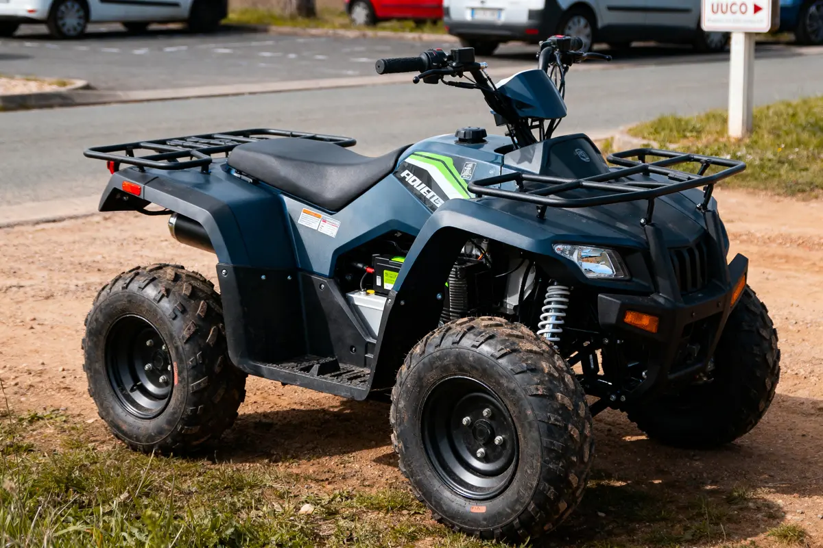 Quad noir garé sur un chemin de terre, avec véhicules et panneaux en arrière-plan.