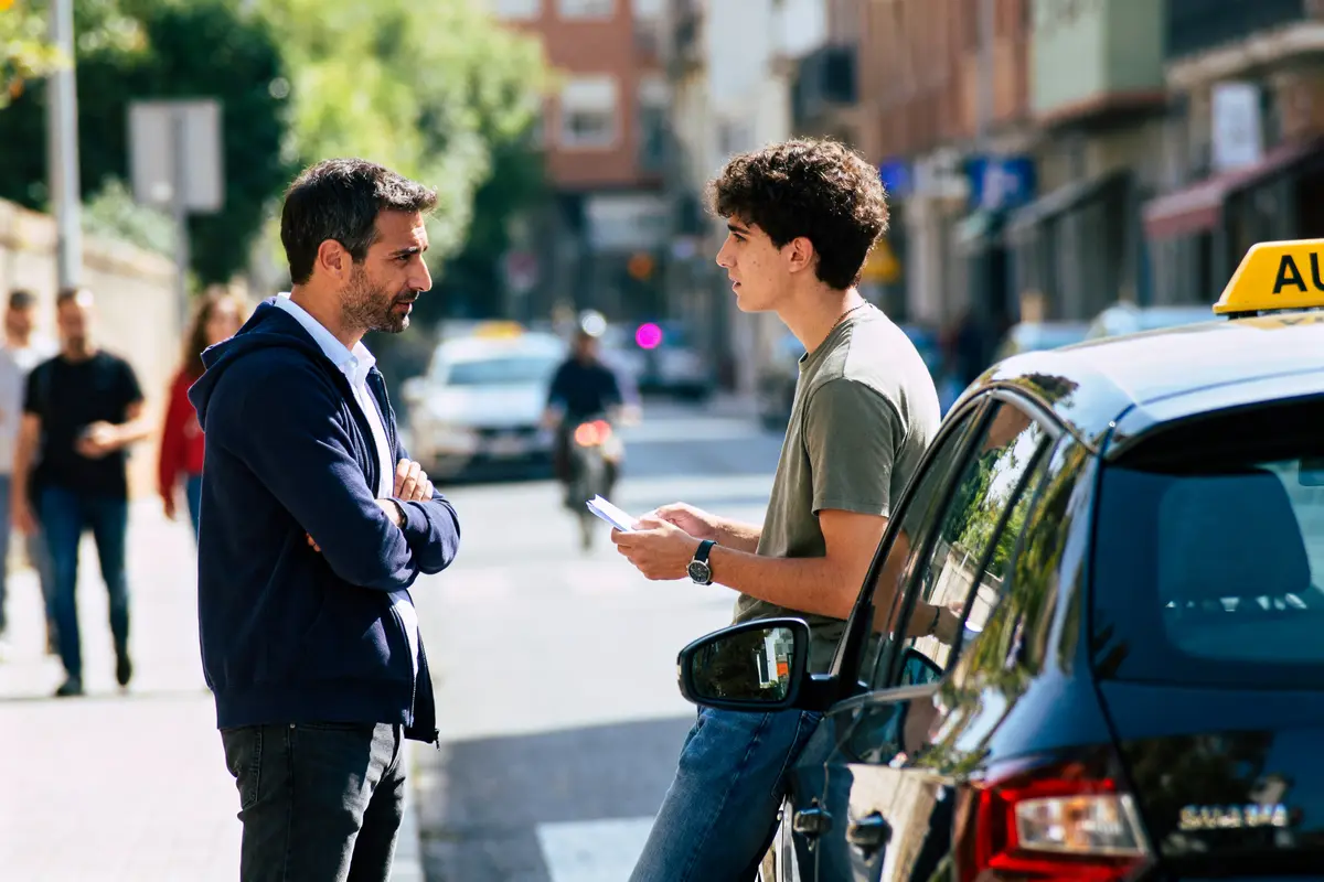 Deux hommes discutant à côté d'un taxi dans une rue ensoleillée. Autres passants flous en arrière-plan.
