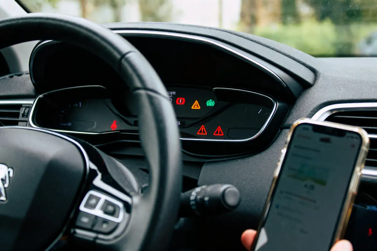 Volant de voiture, tableau de bord avec voyants allumés, personne tenant un smartphone en main.