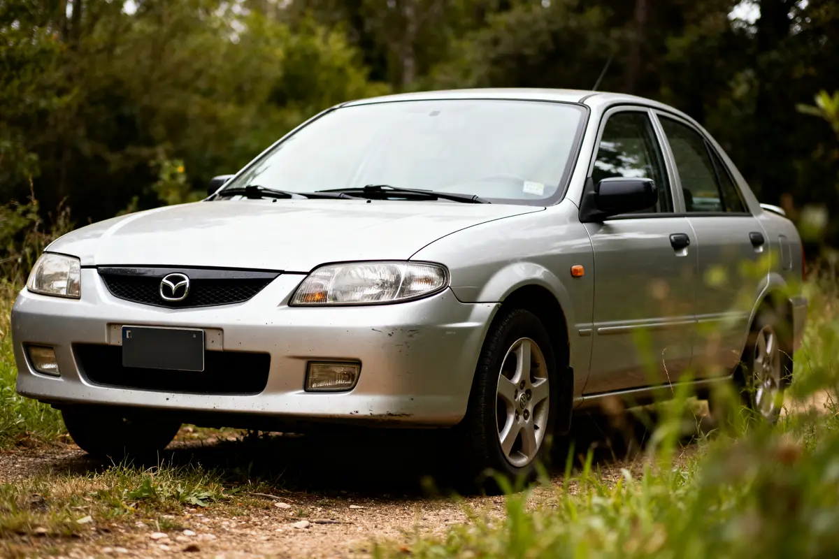 Mazda 323 Protegé argentée garée sur un chemin forestier verdoyant en journée