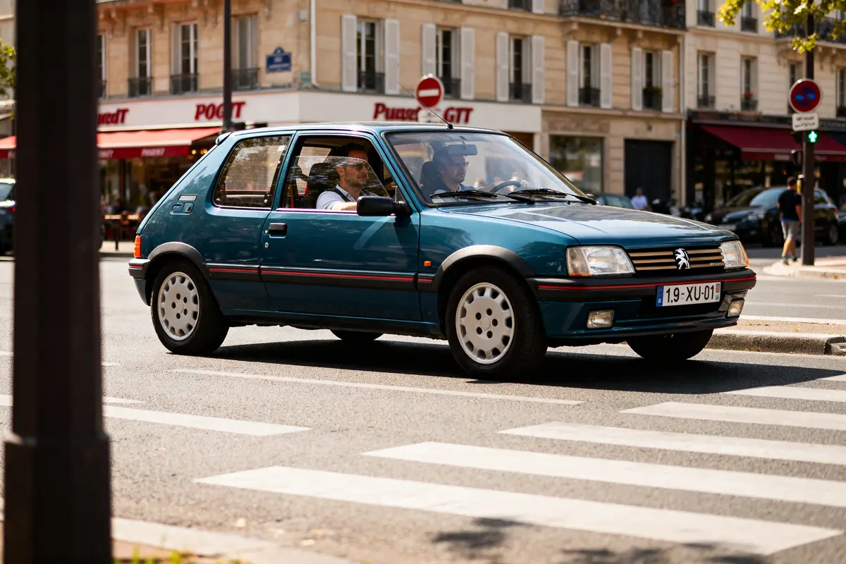 Voiture Peugeot 205 GTI automatic bleue roulant dans une rue urbaine animée